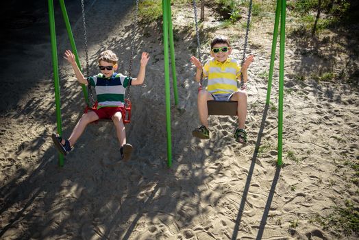 Two little kid boys having fun with swing on outdoor playground. Children, best friends and siblings swinging on warm sunny spring or autumn day. Active leisure with kids. Casual boy fashion.