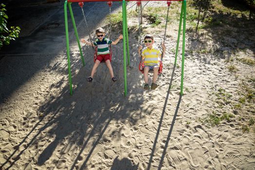 Two little kid boys having fun with swing on outdoor playground. Children, best friends and siblings swinging on warm sunny spring or autumn day. Active leisure with kids. Casual boy fashion.