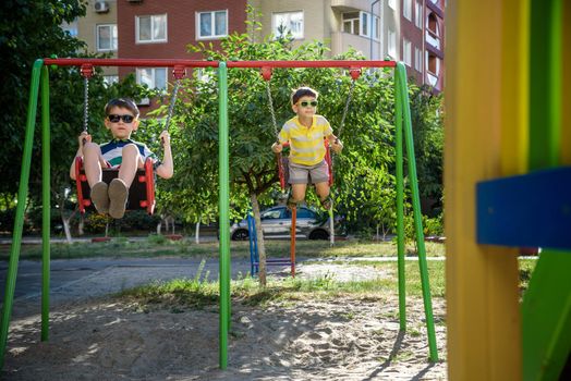 Two little kid boys having fun with swing on outdoor playground. Children, best friends and siblings swinging on warm sunny spring or autumn day. Active leisure with kids. Casual boy fashion.