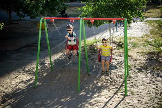 Two little kid boys having fun with swing on outdoor playground. Children, best friends and siblings swinging on warm sunny spring or autumn day. Active leisure with kids. Casual boy fashion.