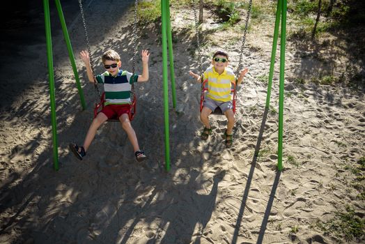 Two little kid boys having fun with swing on outdoor playground. Children, best friends and siblings swinging on warm sunny spring or autumn day. Active leisure with kids. Casual boy fashion.