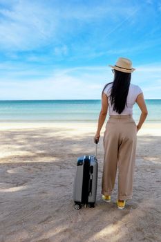 Beautiful young woman wear T-shirt, long pants and straw hat with a suitcase on a tropical beach. Blue sea in the background. Happy travel women standing with travel bag relax on beach. Holiday travel vacation summer