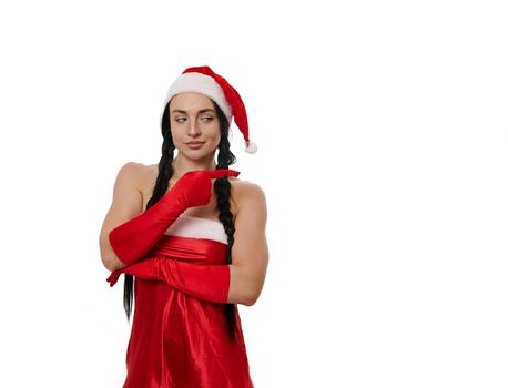 Beautiful sexy dark-haired woman in a carnival costume of Santa Claus smiling cute, looking mysteriously aside and pointing with finger on a copy space of a white wall background. Christmas concept