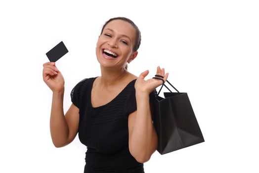 Portrait of young African American mixed race woman smiling with beautiful toothy smile holding shopping bags and credit or discount card in hands, looking at camera standing against white background