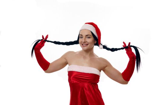 Attractive sexy dark-haired woman in a carnival costume of Santa Claus smiling cute looking at the camera and holding her pigtails to the side, isolated over white background with copy space for ads