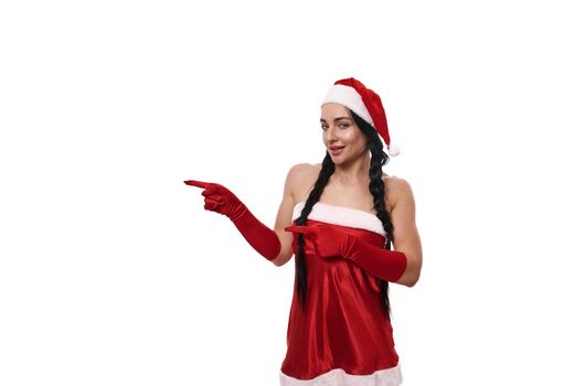 Attractive woman in a Santa Claus carnival costume smiling cute, looking mysteriously at camera and pointing with finger on a copy space, isolated on white background. Christmas advertisement concept