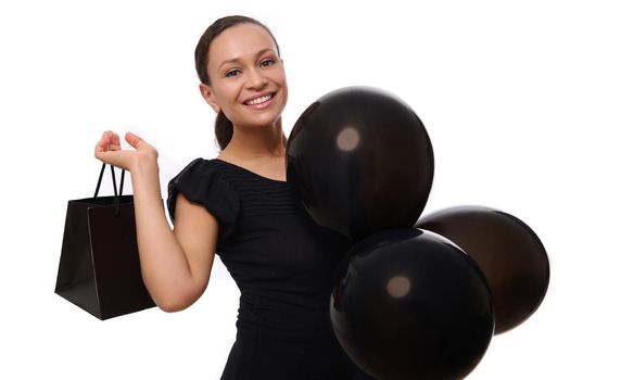 Black Friday concept with attractive mixed race woman with beautiful toothy smile, dressed in black looks at the camera stands with black air balloons and shopping bag, white background, copy space.
