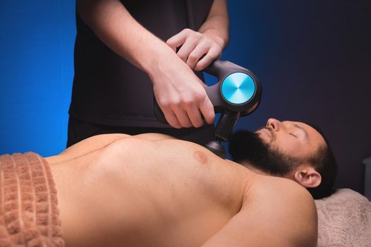 Professional massage of the chest and pectoral muscles with a percussion shock wave massager in a professional office with subdued light.