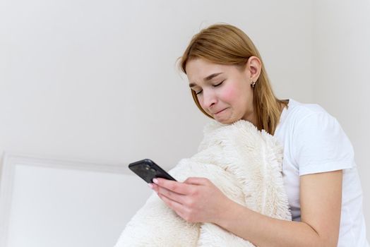 Portrait of a beautiful young woman with a depressed expression, sitting on the bed, holding her phone. Cyber bullying, smartphone addiction and depression, breakup, sad news.