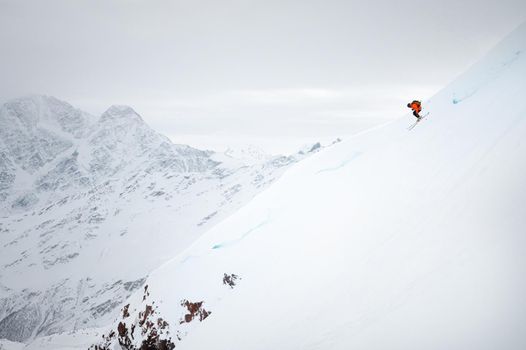 A male skier quickly skis down a snow-covered glacier, against the backdrop of high mountains and a cloudy sky