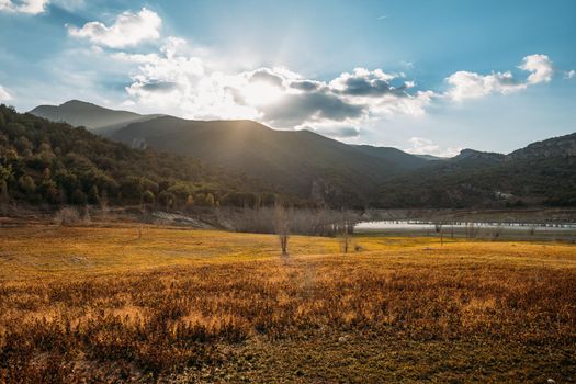 Dawn in the valley in autumn season with orange field and mountains around. Sun shines through the clouds on the blue sky.