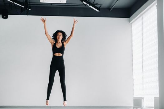 A happy woman in black sportswear makes a jump up on a white background. Girl jumping raises her hands up in the gym.