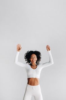 A happy woman in white sportswear bounces on a white background. The girl, jumping, raised her hands up in the gym.