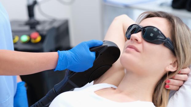 Woman client in protective glasses having laser hair removal of armpit in beauty salon. Body care concept