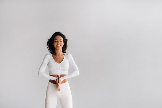 a female yogi in white clothes stands with her hands clasped at the bottom meditating in the yoga hall.