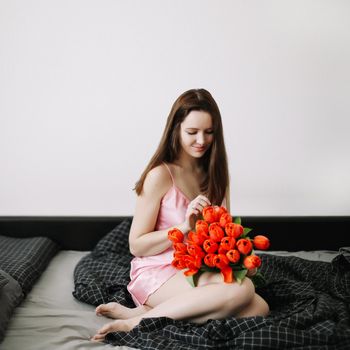 Young beautiful woman sitting in bed and holding a bouquet of red tulips.