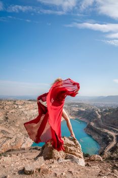 Rear view of a beautiful sensual woman in a red long dress posing on a rock high above the lake in the afternoon. Against the background of the blue sky and the lake in the shape of a heart.