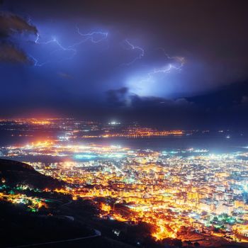 Night lights of the coastal city from the height. Lightning strike on the cloudy dark sky. Fantastic summer night scene.