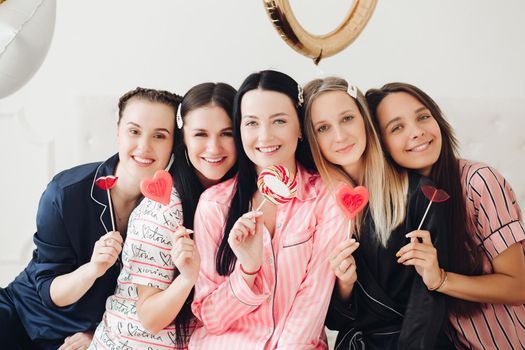 Portrait of happy young female friend posing with colorful sweet candy looking at camera medium shot. Group of smiling girl enjoying pajamas party celebrating holiday having fun together