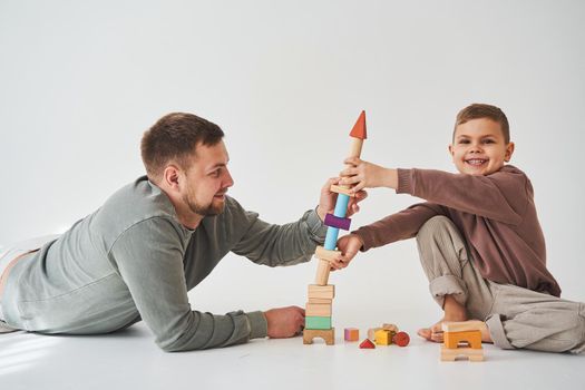 Caring dad helps his son to play on the floor on white background. Father and child build tower of colorful wooden bricks and have fun together