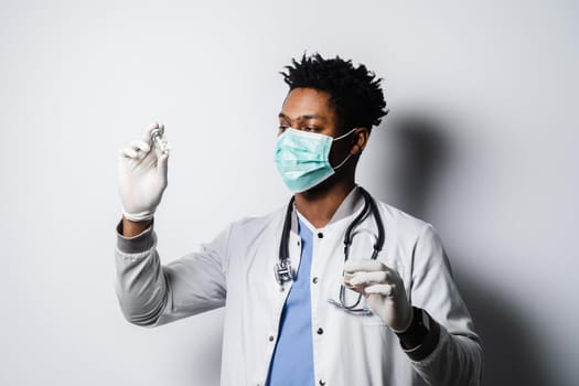 Black doctor in medical mask is holding vaccine on white background. Handsome African doctor with coronavirus covid-19 vaccine