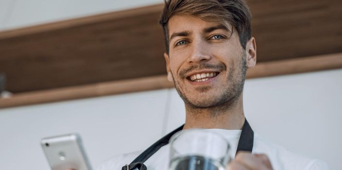 close up. smiling young man with a smartphone standing in the kitchen.