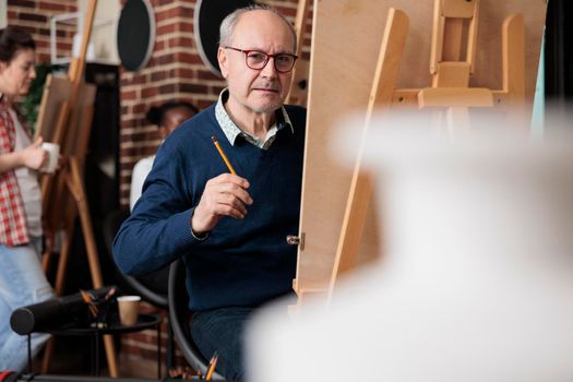 Portrait of senior man student sitting in art class in front on canvas drawing vase model using graphic pencil. Diverse team working at sketching technique developing artistic skill. Creative concept