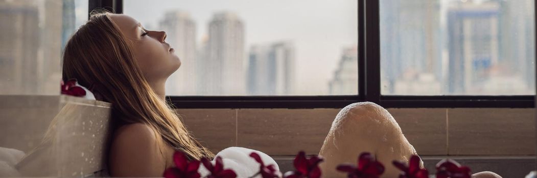 Young woman sits in a bath with foam and frangipani flowers against the background of a panoramic window overlooking the skyscrapers and a big evening city. BANNER long format