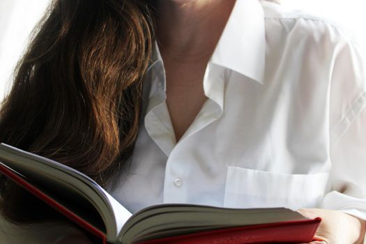 A beautiful girl in a white shirt and long dark hair holds a red book in her hands..