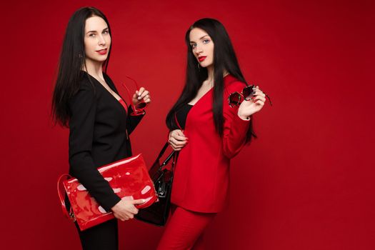 Side view of two brunettes wearing black and red smart suits and glasses standing back to back on isolated background. Stylish women posing and looking at camera in studio. Concept of glamour.