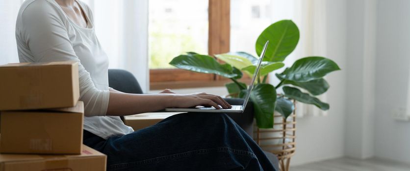 Startup small business owner working with computer at workplace. Freelance woman seller checking orders from laptop. Packing goods for delivery to customer. Online selling. E-commerce.