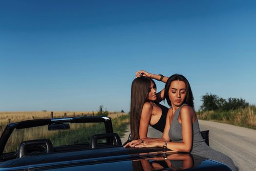 Young women at a photo shoot. Girls gladly posing next to a black convertible against the sky. Summer walk.