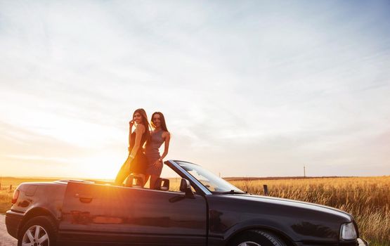 Young two women at a photo shoot. Girls gladly posing next to a black car against the sky on a fantastic sunset.