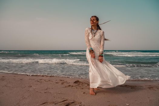 Model in boho style in a white long dress and silver jewelry on the beach. Her hair is braided, and there are many bracelets on her arms