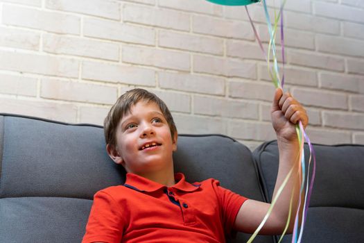a happy European preschool boy is sitting on the sofa and smiling. The boy holds balloons inflated with helium by the ribbons. Birthday Celebration
