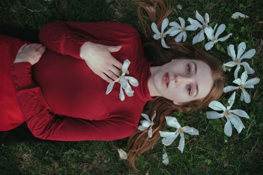 a blonde girl in red lies in the spring on the grass surrounded by magnolia flowers