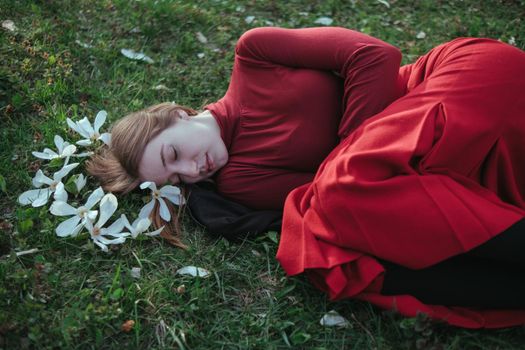 a blonde girl in red lies in the spring on the grass surrounded by magnolia flowers