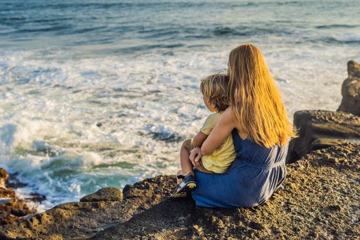 Mom and son are sitting on a rock and looking at the sea. Portrait travel tourists - mom with kids. Positive human emotions, active lifestyles. Happy young family on sea beach.