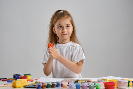 Cute little brunette female artist with a ponytail, in a white t-shirt, is sitting at a table with a whatman and a lot of multi-colored paints and painting her hands. Art studio. Isolated on white background. Drawing process. Medium close-up.