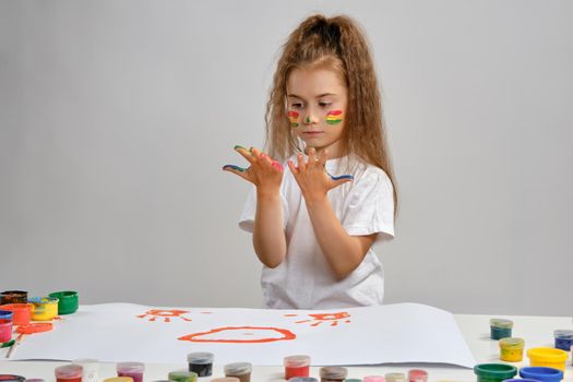 Sweet little brunette female artist with a ponytail, in a white t-shirt, is sitting at a table with a whatman and a lot of multi-colored paints on it and looking at her painted palms. Art studio. Isolated on white background. Drawing process. Medium close-up.
