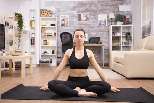 Young woman with keeping eyes open while sitting on the mat in living room doing yoga.