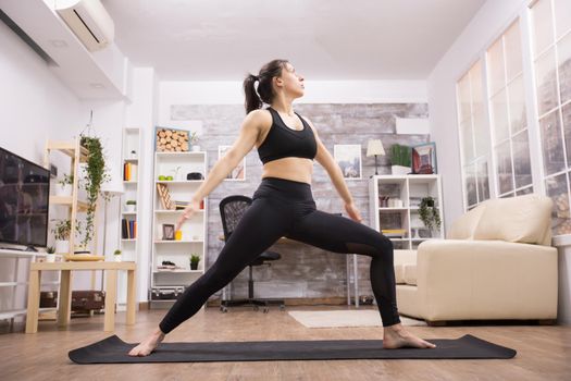 Beautiful brunette standing in warrior yoga position wearing black sportswear in living room.