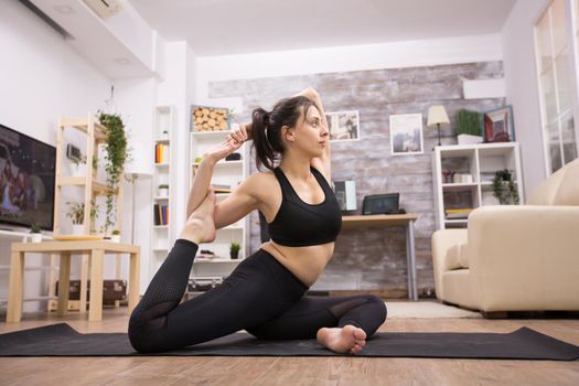 Pretty young woman in black sportswear doing one legged king pigeon yoga pose in living room.