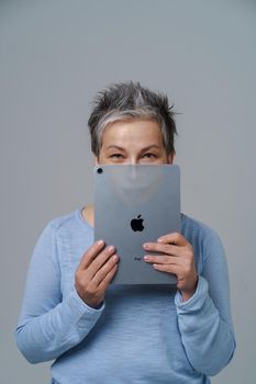 Mature grey haired woman hide hide behind apple ipad posting fake information or bad comments social media with drown evil face or alter ego on tablet isolated on white. Kyiv, Ukraine, May 13, 2022