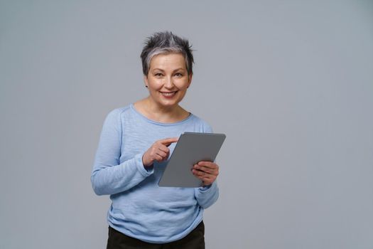 Smiling mature grey haired woman 50s holding digital tablet working or checking on social media. Pretty woman in 50s in blue blouse isolated on white. Older people and technologies. Toned image.