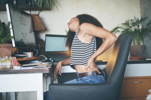 Shot of a young african woman sitting alone and suffering from back ache while using her computer to work from home stock photo