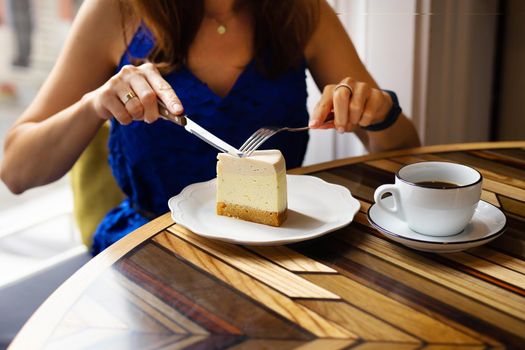 Young girl cuts a piece of cream cheesecake, on the background of a cup of coffee, close-up