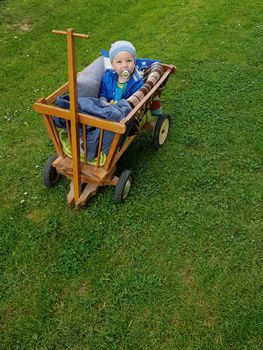 Small child sitting in a wooden wagon coverd with blankets surrounded with green grass. Looking straight in to a camera.