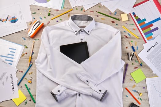 Top view flat lay white shirt with tablet device. Stapler with paper clips on wooden desk.