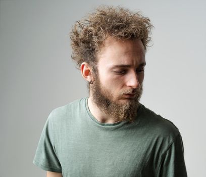 Pensive young handsome bearded man with wild curly hair, bright blue eyes looking down isolated on white background. Young thinking man in green t shirt on white.